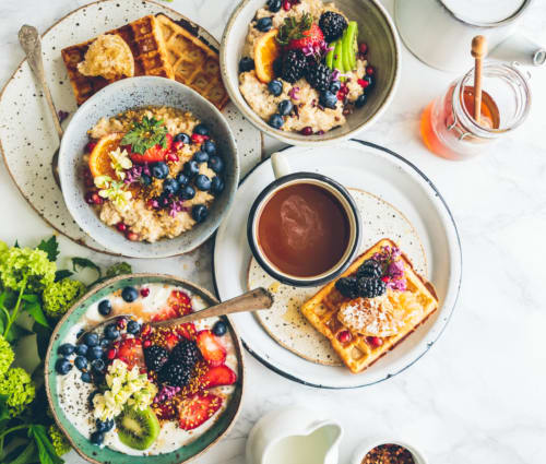 A breakfast spread with bowls of oatmeal topped with fruits and berries, accompanied by waffles and a cup of coffee.