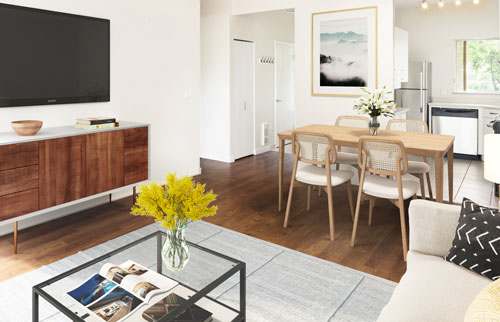 Image of a staged suite, the view is from the living room looking over the coffee table back to the dining room. The furniture is a contemporary adaptation of the mid-century modern style.  In this image, the TV mounted on the wall is visible.