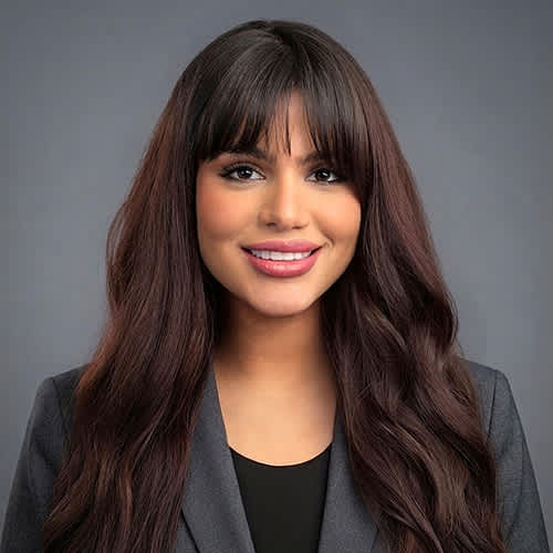 A woman with long brown hair and bangs wearing a grey blazer over a black top.