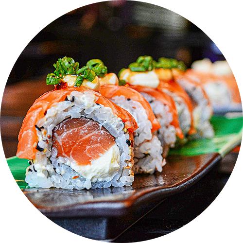 A sushi roll with salmon and cream cheese on top is placed on a dark plate.
