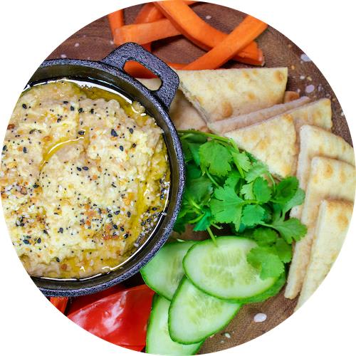 A bowl of hummus topped with olive oil and black pepper sits on a plate with pita bread, cucumber slices, and carrot sticks.