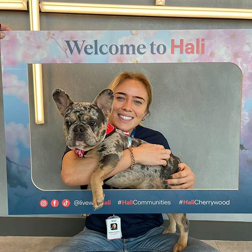 A woman is holding a dog in front of a sign that says "Welcome to Hali".