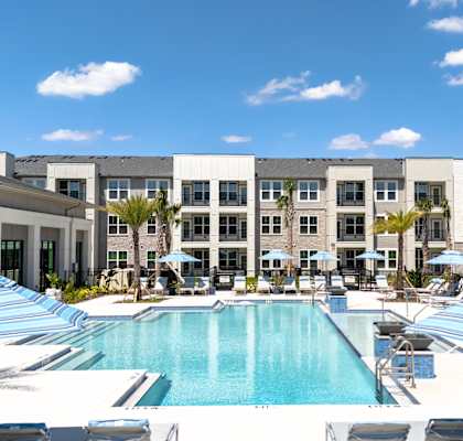 A large swimming pool surrounded by lounge chairs and umbrellas in front of a multi-story building.