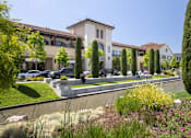 Thumbnail 10 of 13 - A building with a white facade and a brown roof is surrounded by greenery and a small pond.