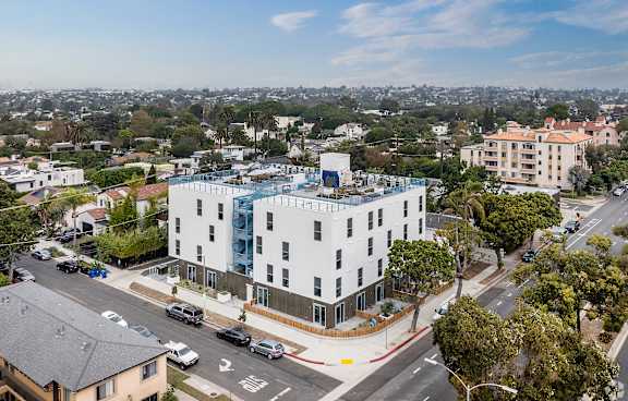 A white building with a blue glass addition sits on a street corner with cars parked on the side.