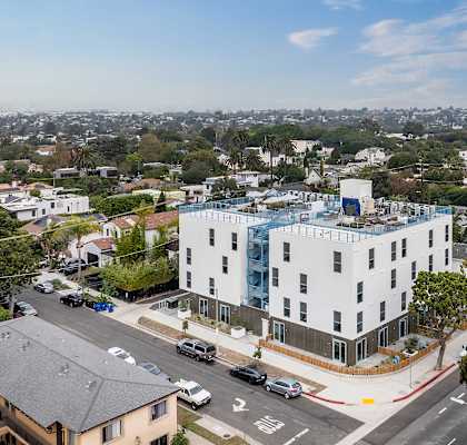A white building with a blue glass addition sits on a street corner with cars parked on the side.