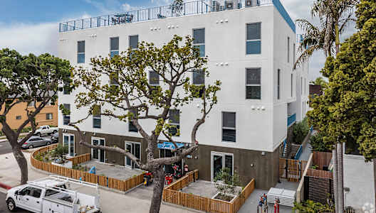 A white building with a balcony on the top floor is surrounded by trees and has a white van parked in front.