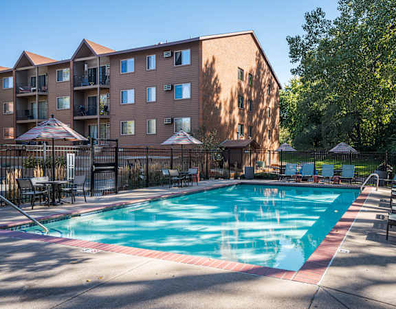 A large swimming pool surrounded by lounge chairs and umbrellas in front of a multi-story apartment building.