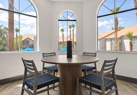 A round wooden table with four chairs in front of a window with a view of a house and trees.