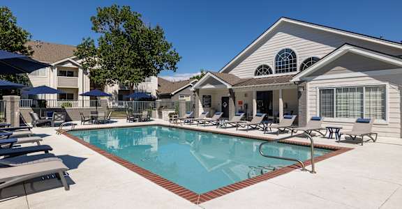 A swimming pool in front of a house with a patio and lounge chairs.
