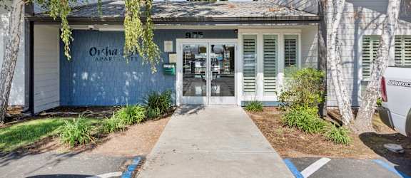 The entrance to a building with the sign "Orchard Apartments" is surrounded by trees and plants.