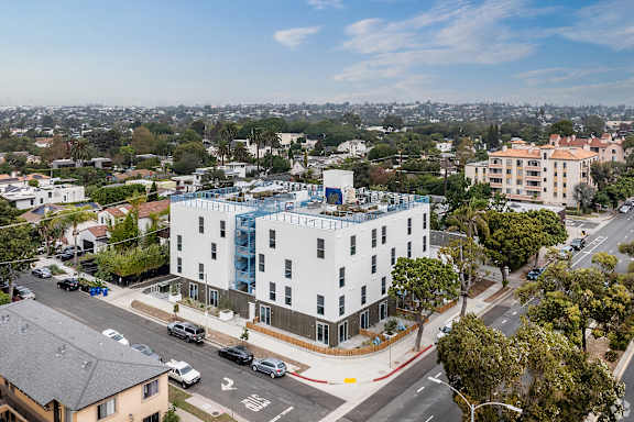 A white building with a blue glass addition sits on a street corner with cars parked on the side.