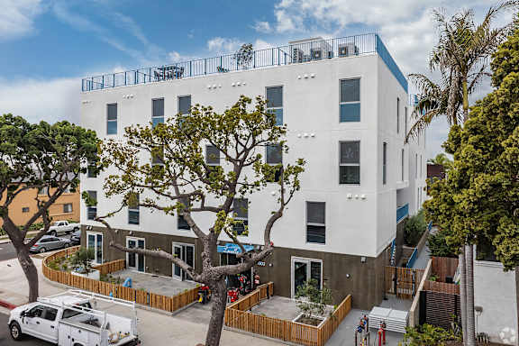 A white building with a balcony on the top floor is surrounded by trees and has a white van parked in front.