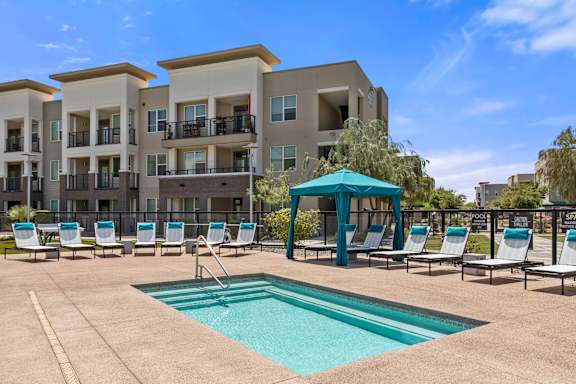 A swimming pool area with sun loungers and a blue umbrella in front of apartment buildings.