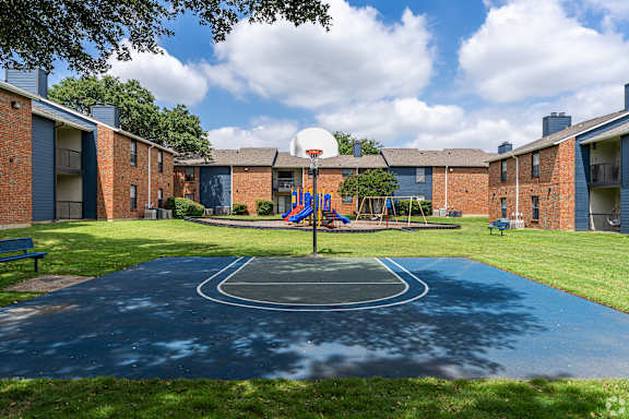 A basketball court in the middle of a grassy area with apartment buildings in the background.