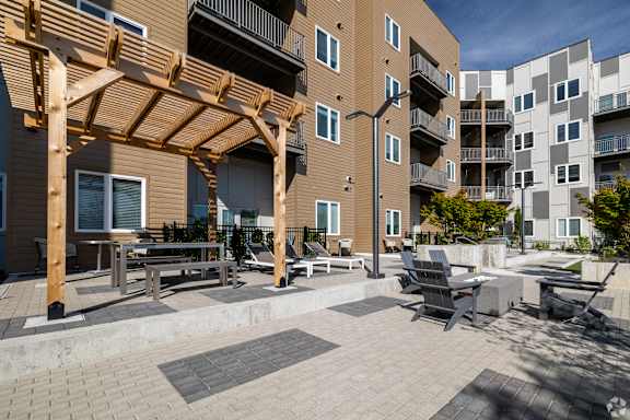 A patio area with a wooden pergola and seating area in front of apartment buildings.
