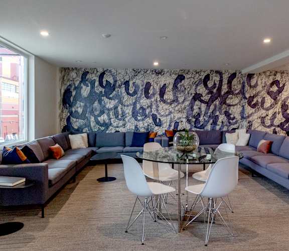 A living room with a large tapestry on the wall and a glass table in the center.