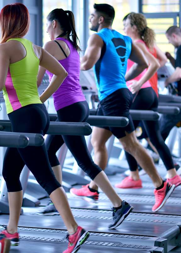 A man in a white t-shirt is running on a treadmill in a gym with other people exercising around him.