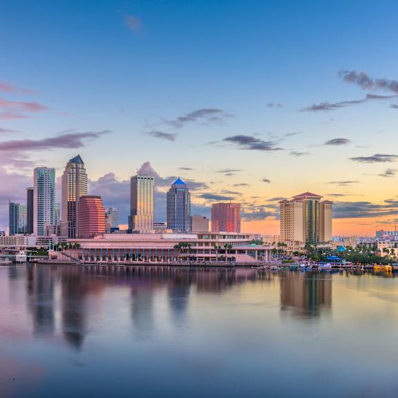 A city skyline at sunset with buildings reflecting in the water.