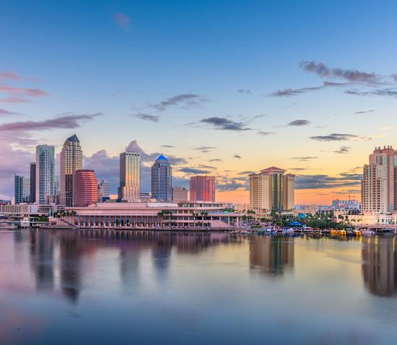 A city skyline at sunset with buildings reflecting in the water.