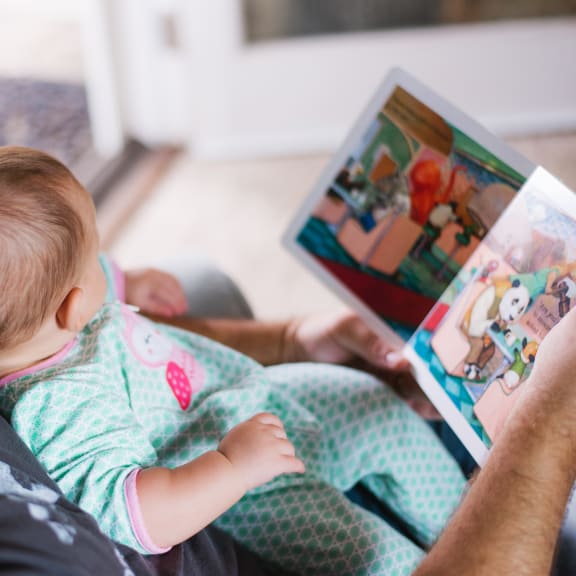 a man sitting on a couch holding a baby and reading a book at King West One, Montana