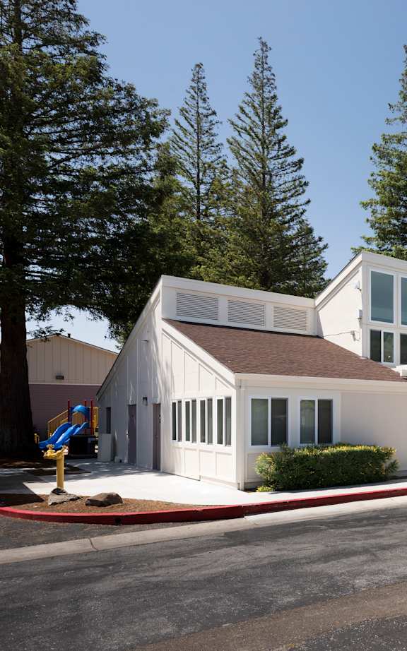 A white building with a brown roof is surrounded by trees.