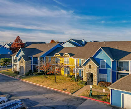 The beautiful exterior of the blue and yellow homes and the green space with grass, trees, and shrubs, with the parking lot at Woodland Park Apartment Homes. 