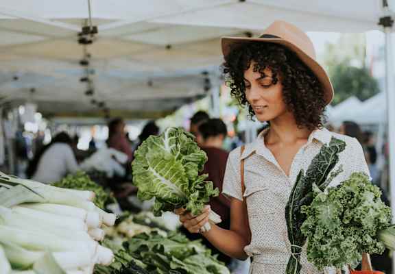 A woman in a hat is holding a bunch of lettuce and other vegetables at a market.
