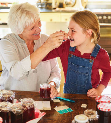 A young girl in a red shirt and blue denim overalls eating some jam from a spoon fed from her grandmother.