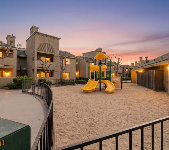 A playground with a yellow slide is surrounded by a sandy area and a fence.