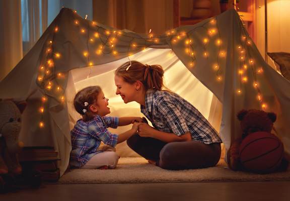 A woman and a child are sitting inside a tent-like structure with lights strung up around it.