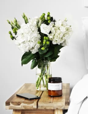Wooded bedside table with a brown candle and glass vase with white and green flowers in it. A white bed is next to the wooded table. 