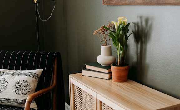 A chair with a striped back and a pillow with circles on it sits next to a wooden table with a vase of flowers, a plant, and a stack of books.