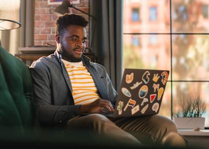 A man in a striped shirt and grey blazer is working on a laptop with a sticker-covered case.