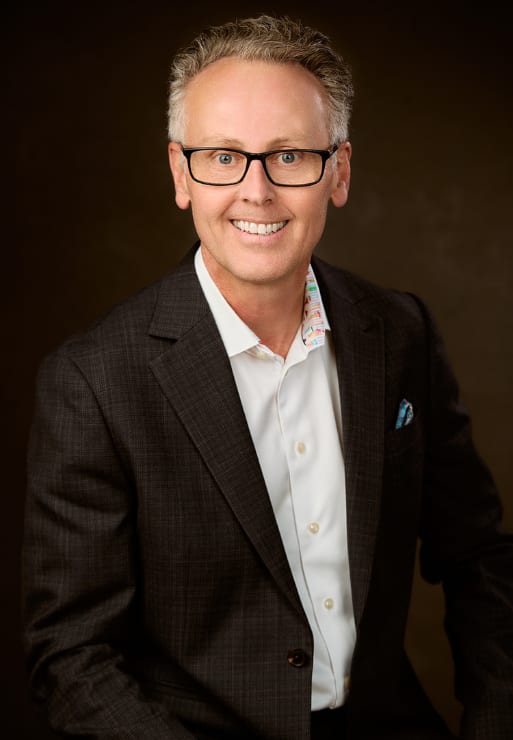 A man with glasses and a blue checkered shirt is wearing a dark blue suit.