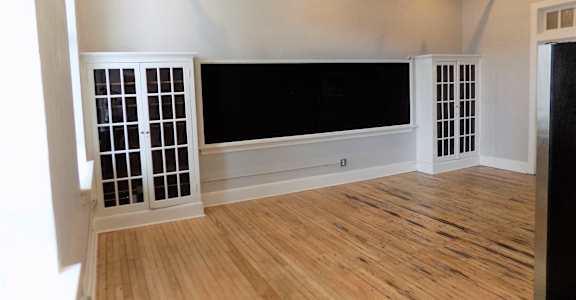 A room with wooden floors and white walls has a large black screen and two windows with white grills.