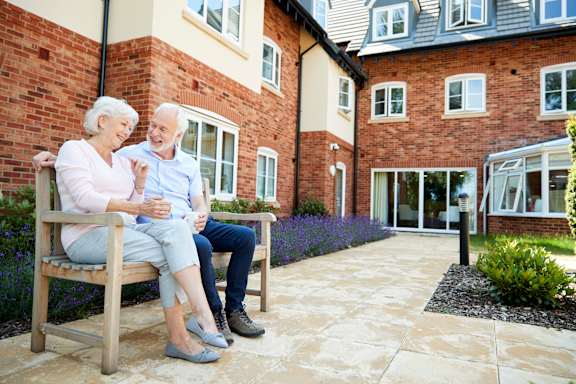 An elderly couple is sitting on a bench outside a brick building.