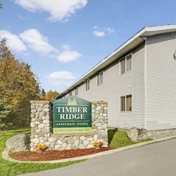 The exterior sign surrounded by rocks and a large tree behind it with the buildings in the background of Timber Ridge Apartment Homes. 