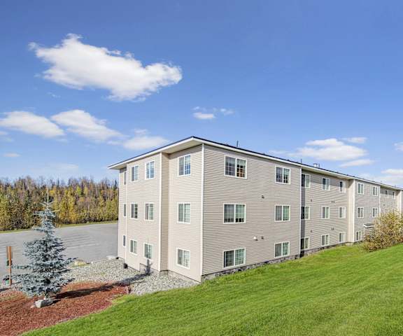 The exterior of the white building with a parking lot and green space with trees at Timber Ridge Apartment Homes. 