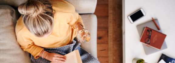 A woman is sitting on a couch with a green blanket, holding a book and a cup of coffee.