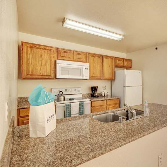 A spacious kitchen with white appliances, kitchen utensils, overhead lighting, light countertops with lots of room, a bar counter with barstools, and large wooden cabinets at Timber Ridge Apartment Homes. 
