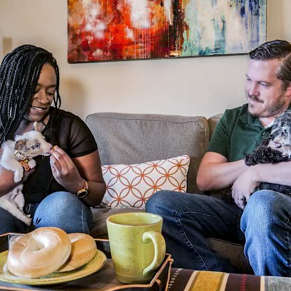 A man and woman sitting on a couch with two dogs in a living room at 3600 Brentwood Apartment Homes.