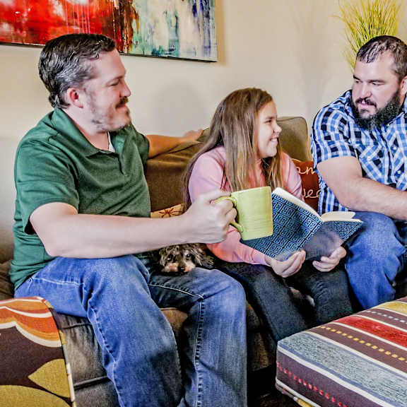 Two dads and their daughter sitting on a couch in a living room reading at Ranchlands Apartment Homes.