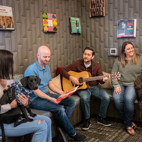 A group of people sitting in a room with a dog and a guitar at Prairie Vista Apartments. 