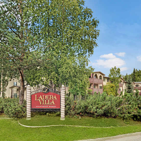 The exterior sign with trees and surrounded by greenery with the buildings in the background of Ladera Villa Apartments. 