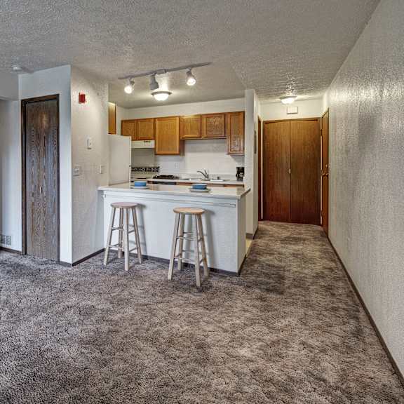 A cozy kitchen with white appliances, blue kitchen utensils, overhead lighting, light countertops with lots of room, a bar counter with wood and gray barstools, and large wooden cabinets at Russian Jack Apartment Homes. 