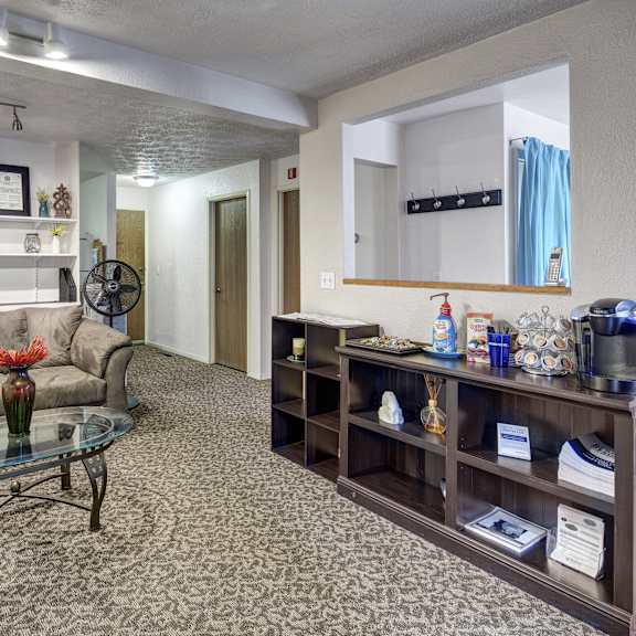 The resident lounge with a plush gray couch, a coffee station with a coffee maker, coffee, and add ons, and a shelf with decorations at Russian Jack Apartment Homes. 