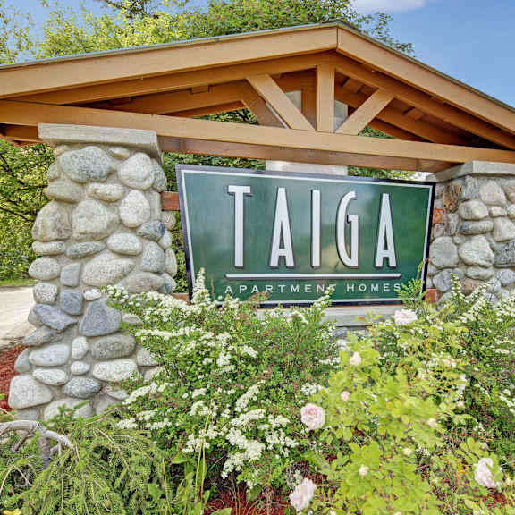 The exterior sign with trees and surrounded by greenery with the buildings in the background of Taiga Apartment Homes. 