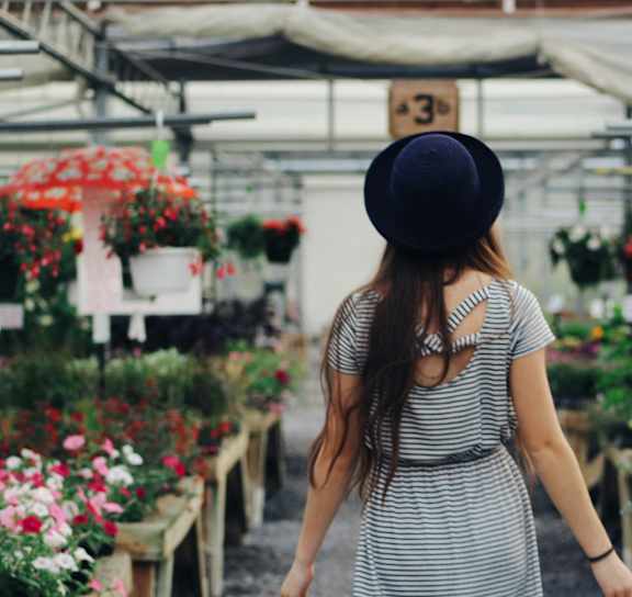 A woman in a striped dress and hat is walking through a greenhouse filled with plants.