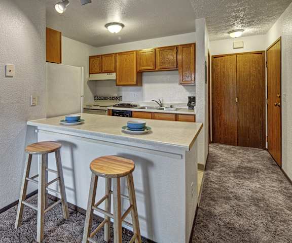 A cozy kitchen with white appliances, blue kitchen utensils, overhead lighting, light countertops with lots of room, a bar counter with wood and gray barstools, and large wooden cabinets at Russian Jack Apartment Homes. 